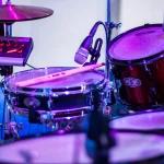 A snare drum and tom fitted with microphones on a stage under purple light, beside a small mixer and headphones